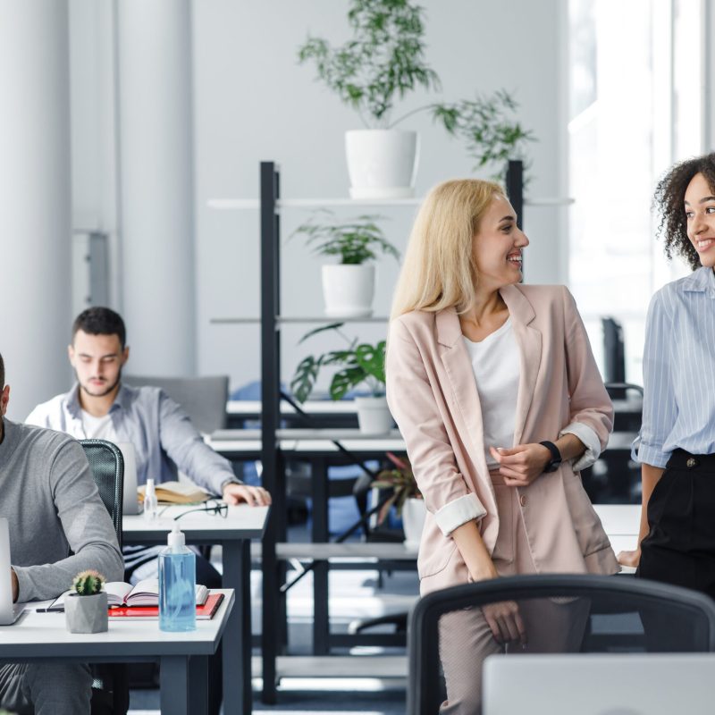 Turning to work, new normal and communicating after lockdown. Happy european and african american women gossip in interior of modern office with colleagues at workplaces during pandemic, free space